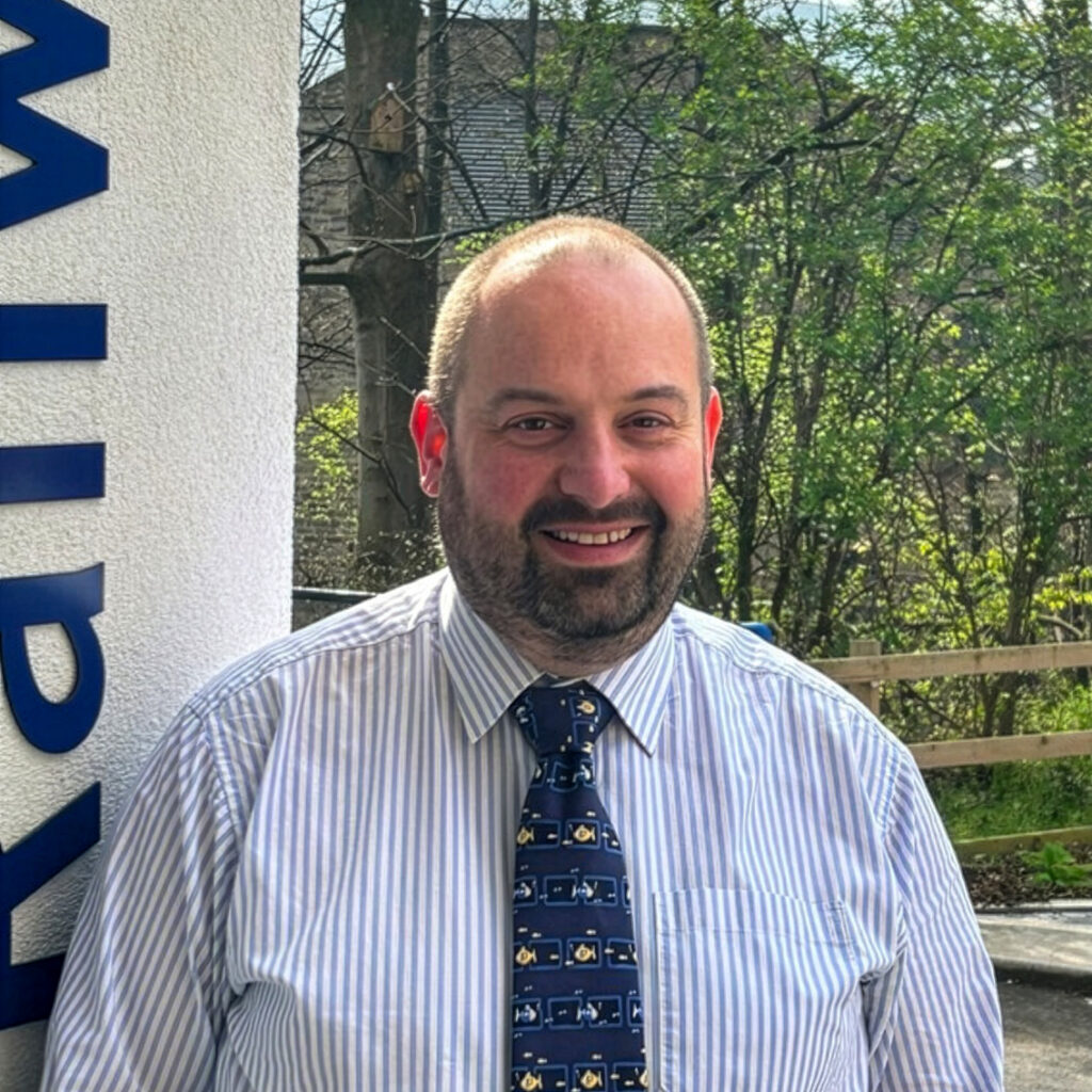Individual in striped shirt and patterned tie stands outdoors near building signage, with trees and wooden fence in the background.