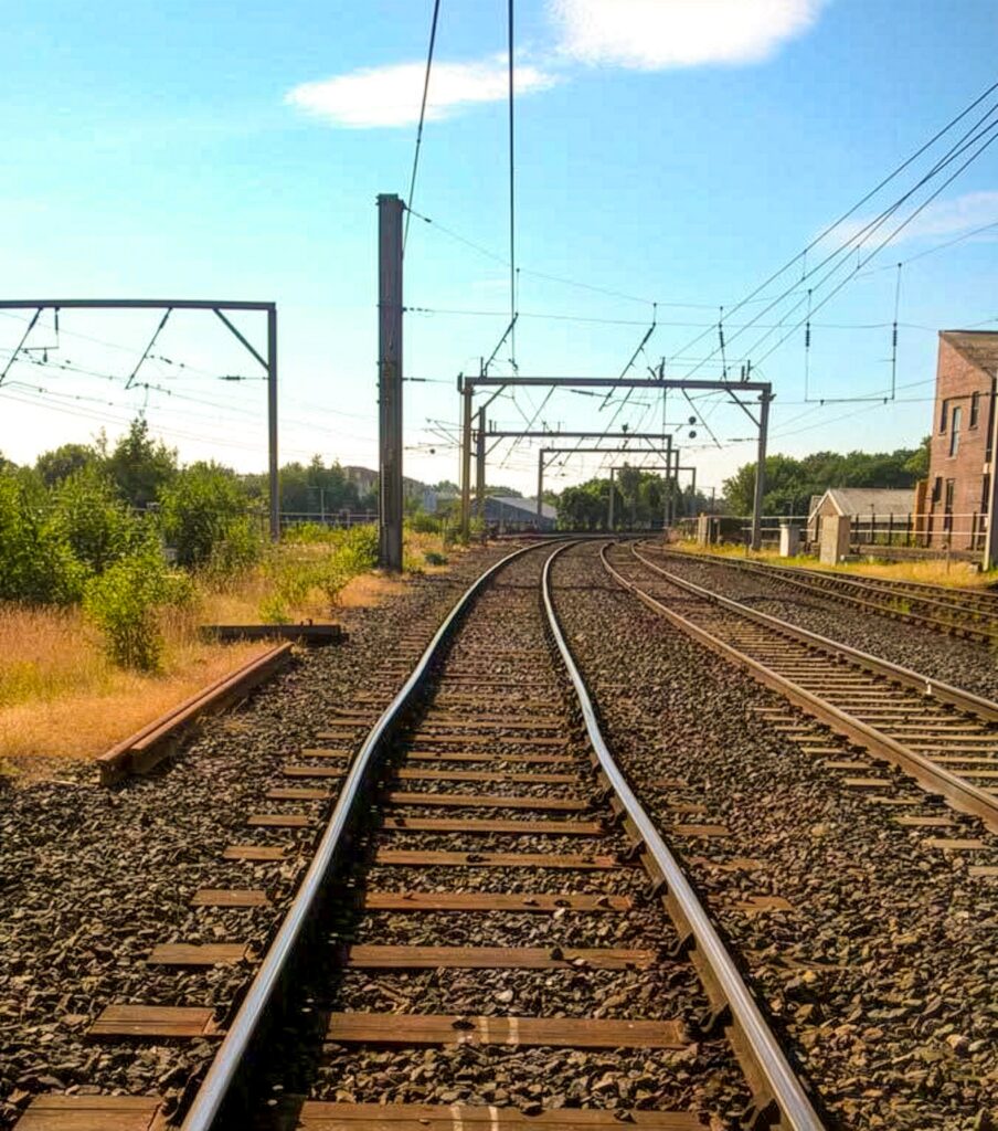 Railway tracks curve and buckle through outdoor landscape with overhead electrification lines, pylons and surrounding vegetation under blue sky.