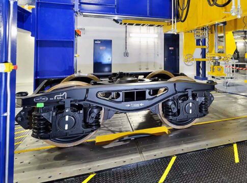 Railway bogie mounted on testing rig inside engineering laboratory, with blue and yellow machinery and industrial equipment visible.